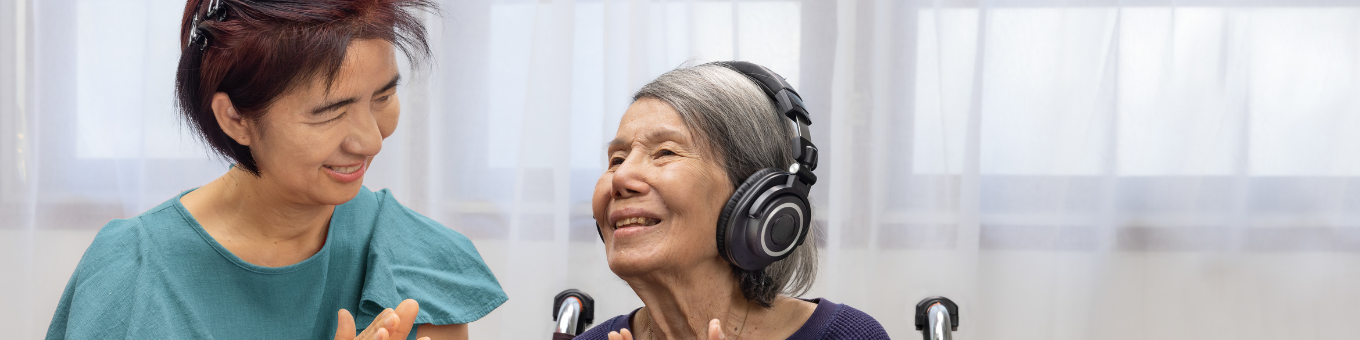 A women with dementia wearing headphone and sitting next to her carer both clapping their hands