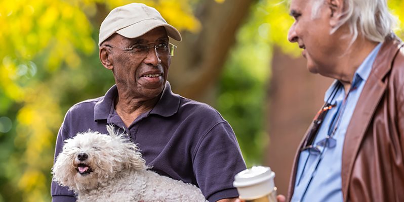 Two men talking, one holding a dog and another holding a coffee cup