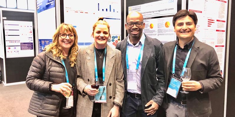 Four HIV researchers wearing lanyards and stood next to posters at a conference